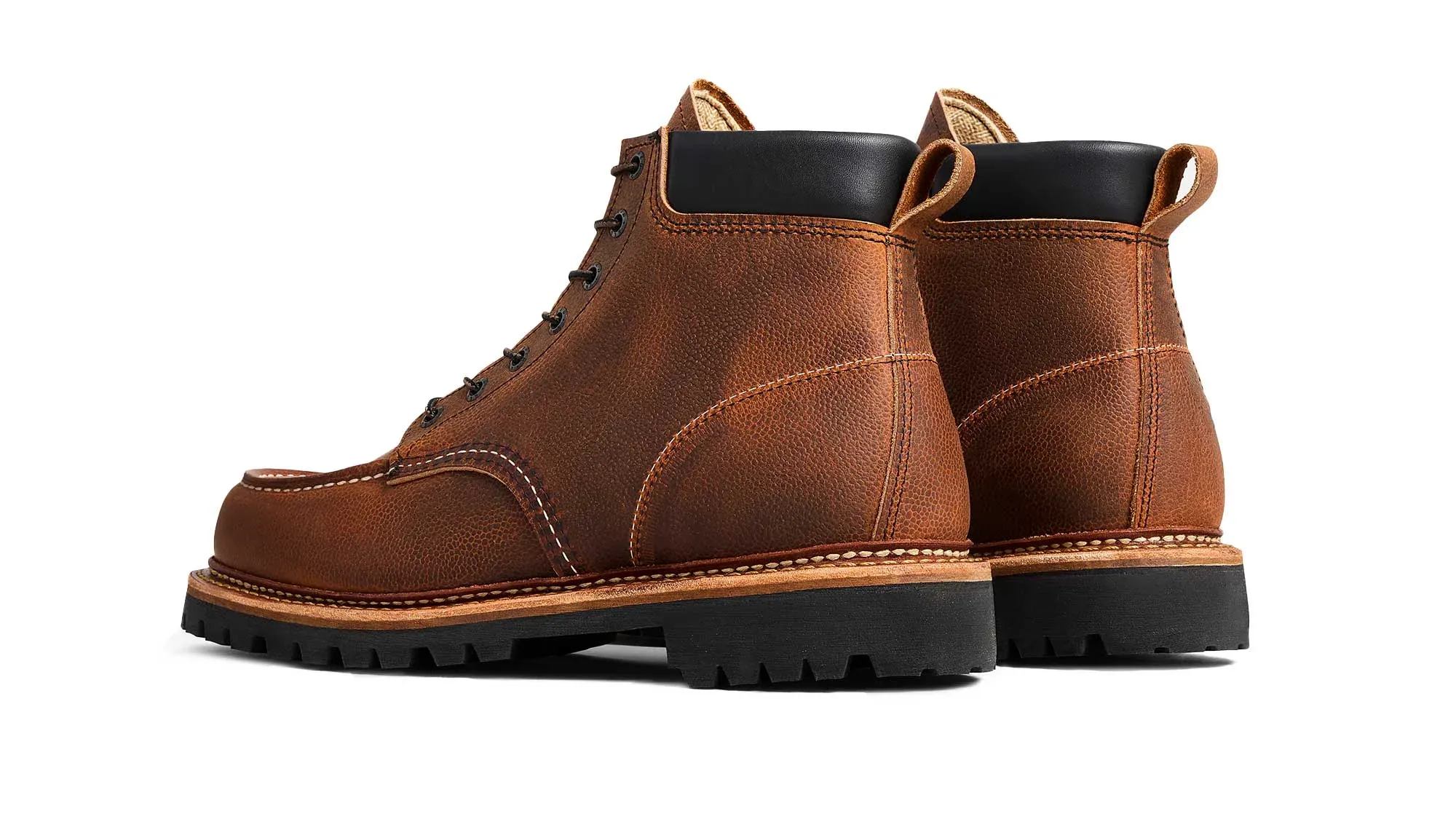 A pair of brown leather moc toe boots on black lug soles, seen from behind at an angle